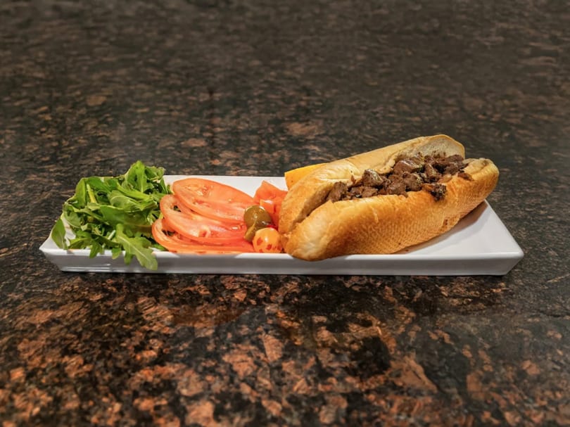 Cheesesteak sandwich with a side of arugula, sliced tomatoes, and pickled peppers on a white plate.