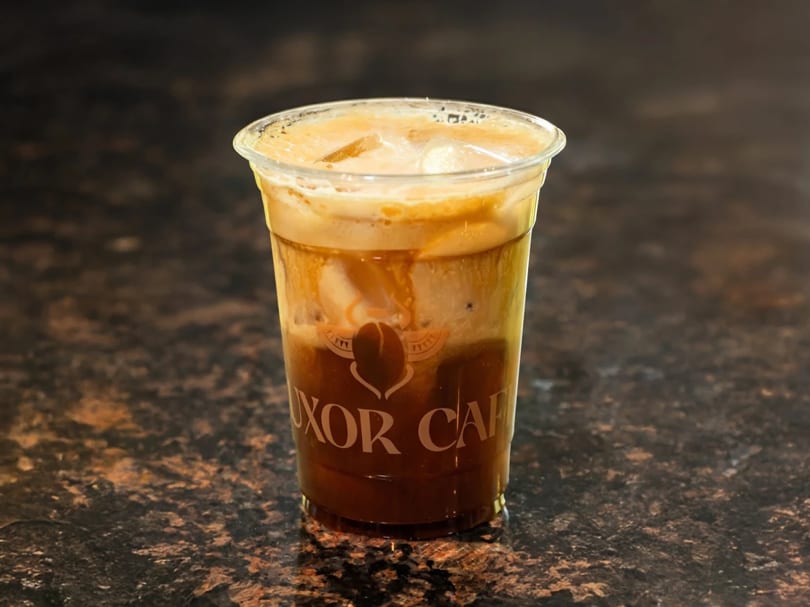 Iced coffee with cream and ice in a clear plastic cup with the Luxor Cafe logo.