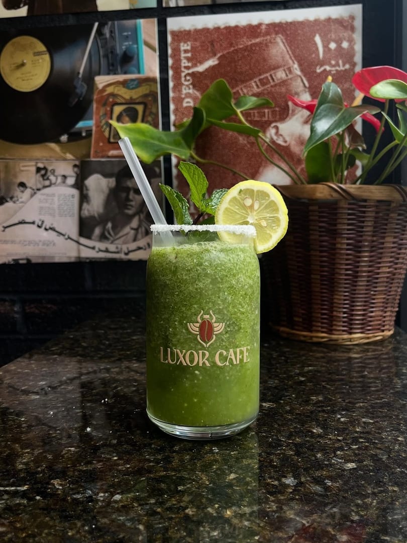 A green Luxor Cafe drink with mint and lemon on a dark counter, with vintage decor in the background.